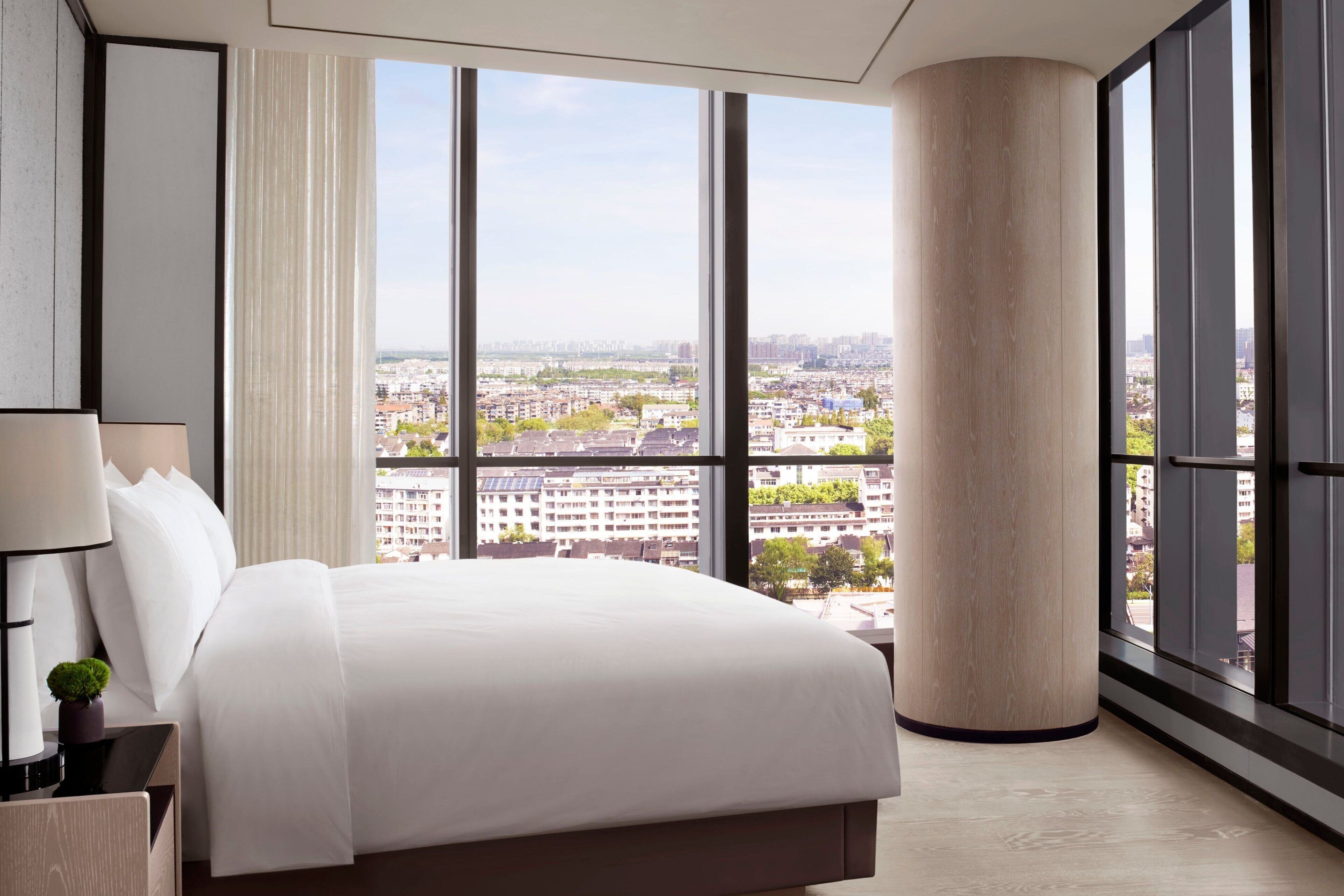 A modern hotel room with a large bed, a view of the city through floor-to-ceiling windows, and a large pillar in the corner.
