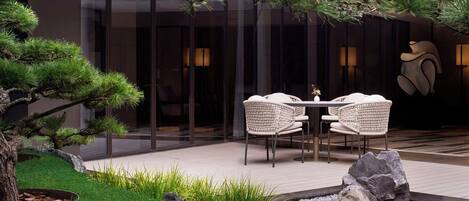 A modern outdoor seating area with a table and chairs, surrounded by a well-maintained garden with a pine tree and rocks.