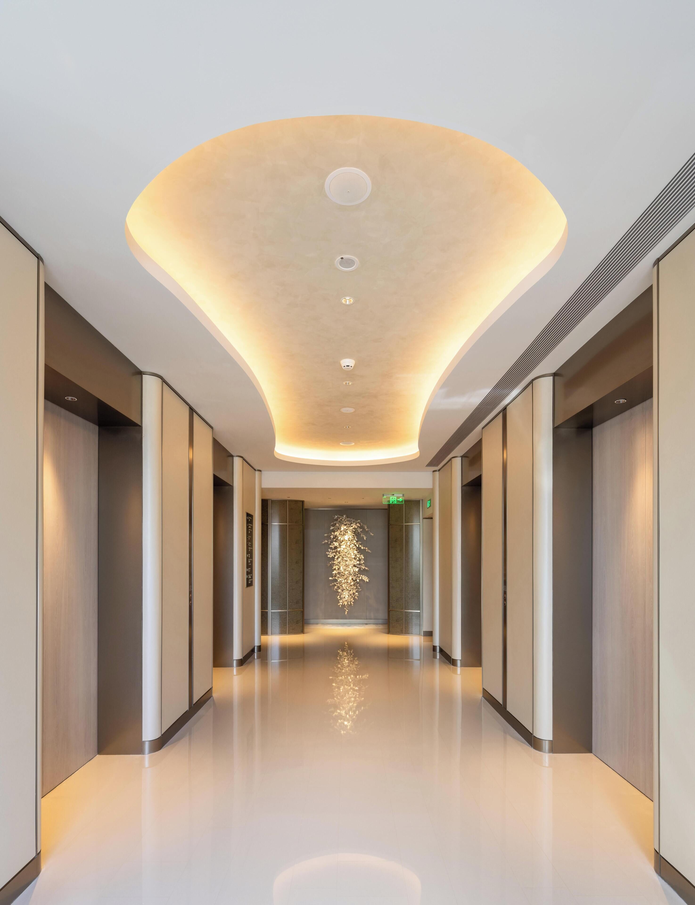 A modern hotel corridor with a curved ceiling, recessed lighting, and a reflective floor.