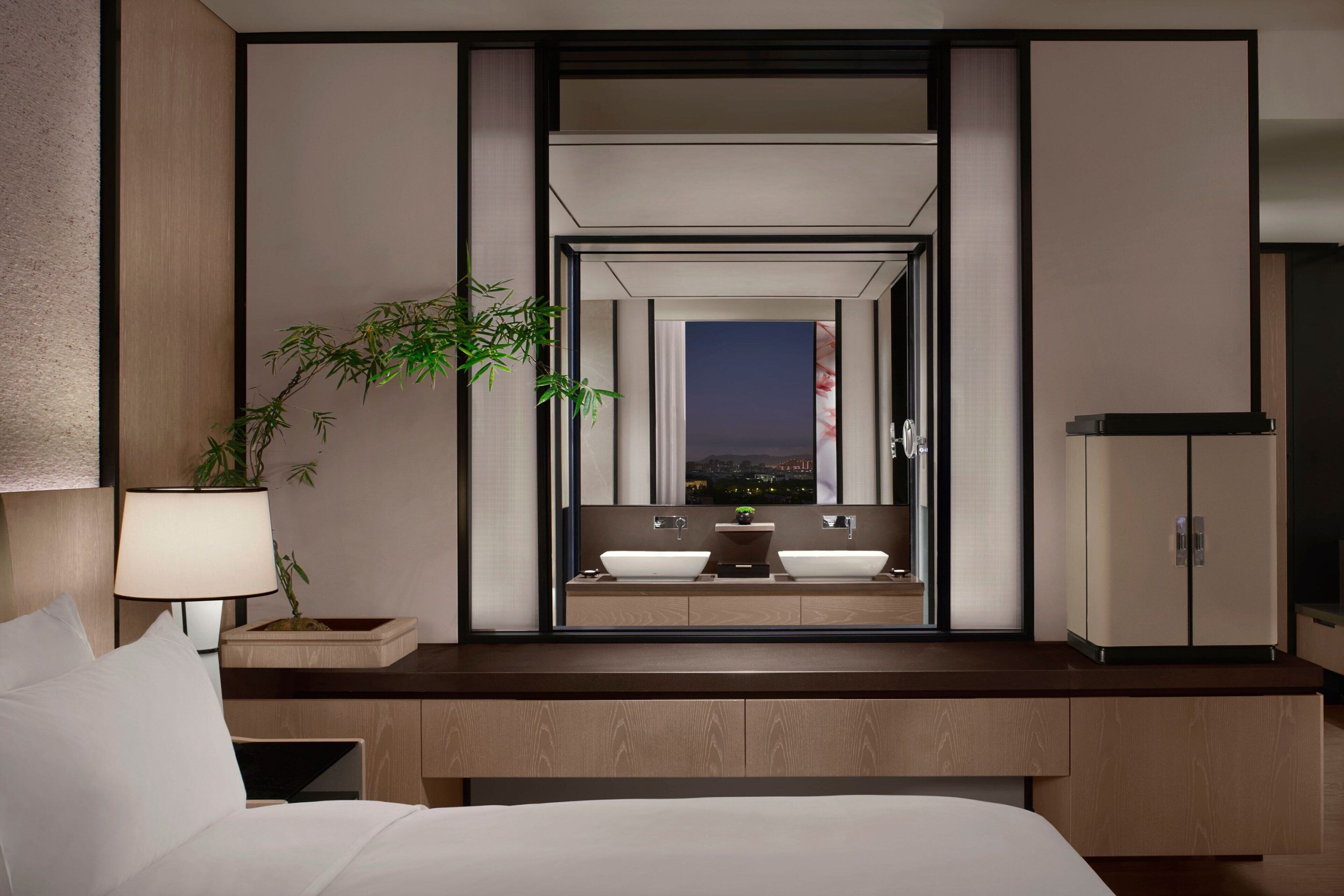 Une chambre d’hôtel avec un lit, une table de chevet avec une lampe, une plante, un grand miroir qui reflète une salle de bain avec deux lavabos, et une vue sur le paysage urbain de nuit.