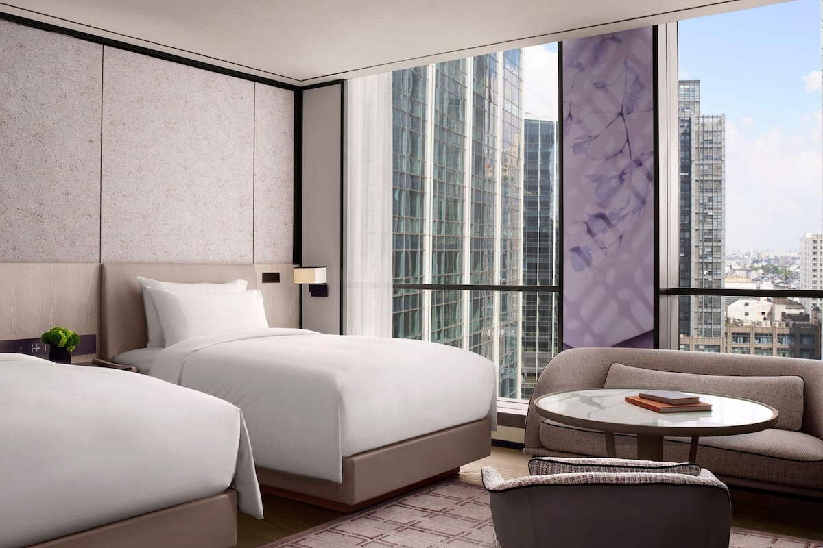 A modern hotel room with two beds, a sofa, a small table, and a large window offering a city view.