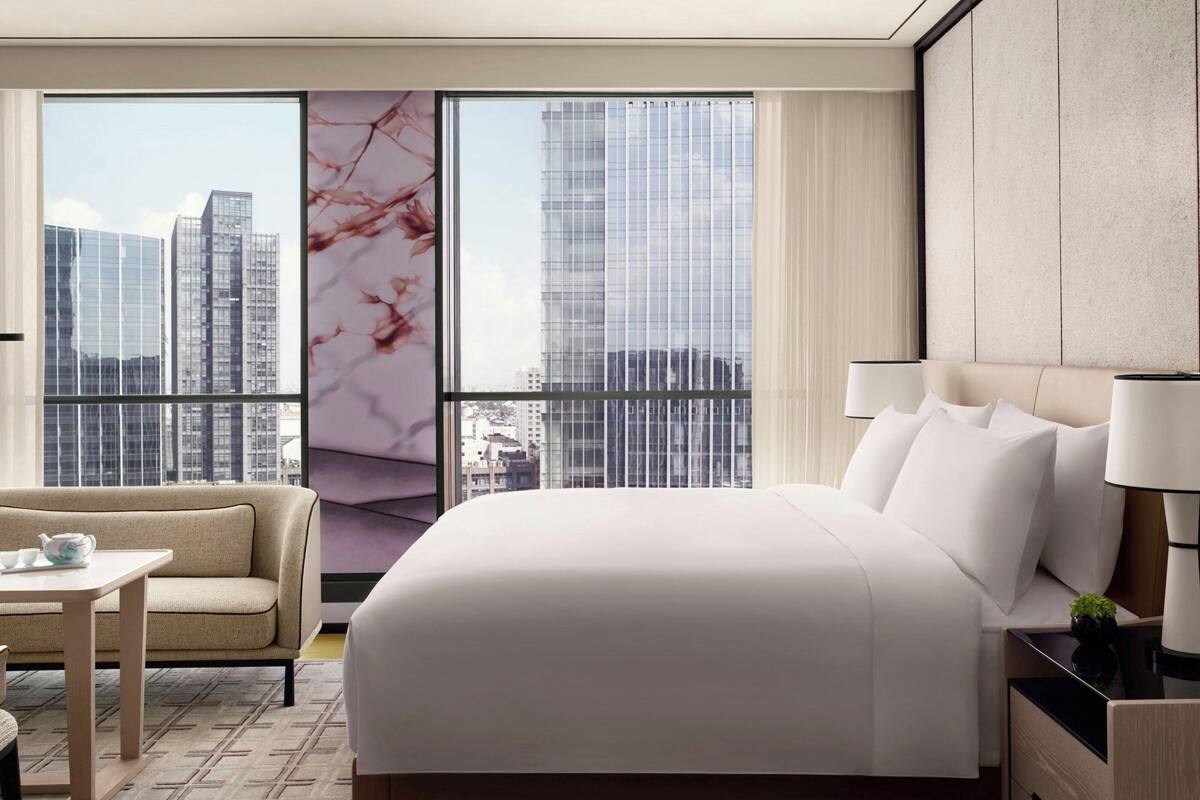 A hotel room with a large bed, a sofa, a small table, and a view of tall buildings through large windows.