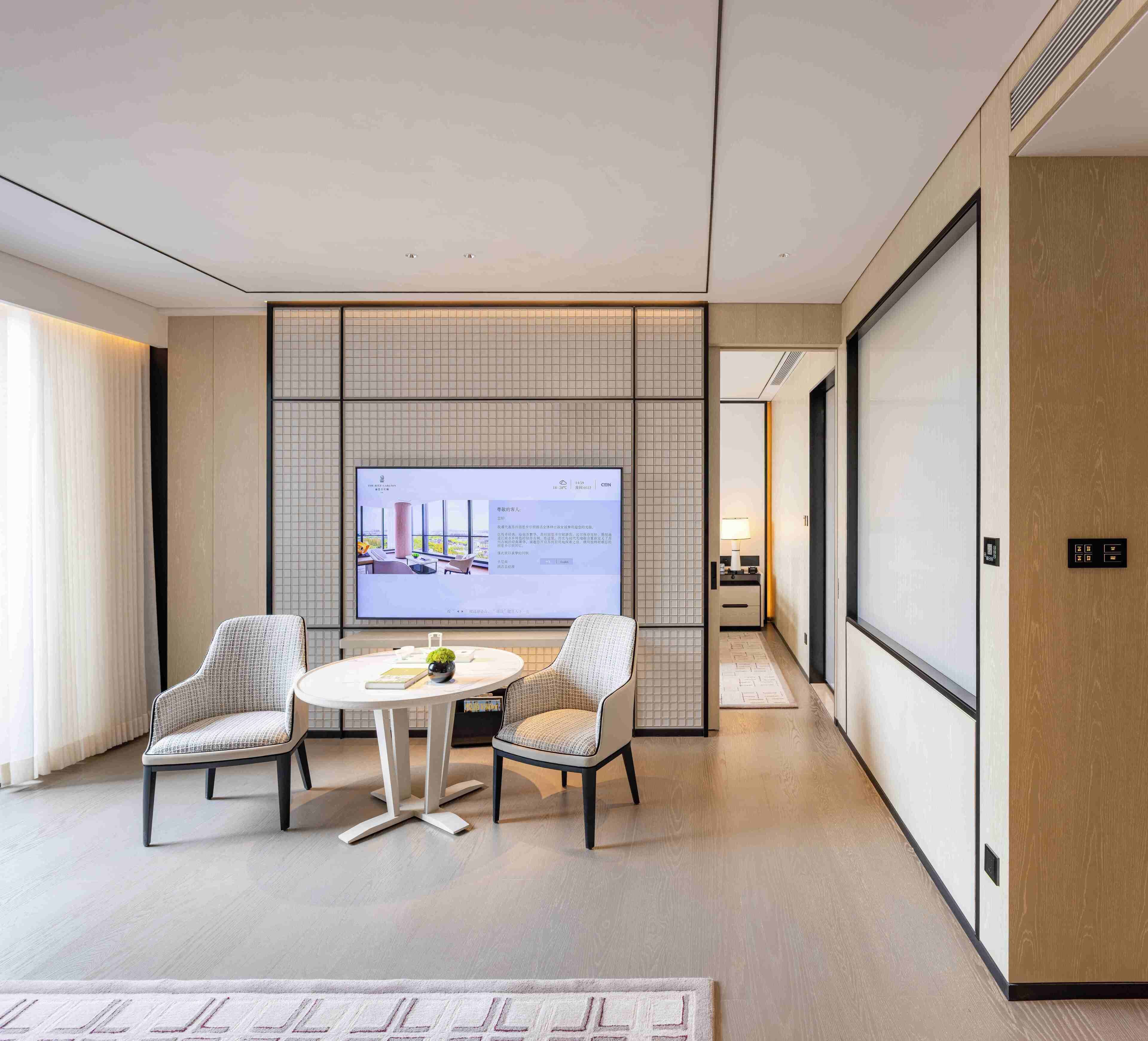 A modern hotel room with a large flat-screen TV, two upholstered chairs, a round table, and a traditional Japanese-style partition.