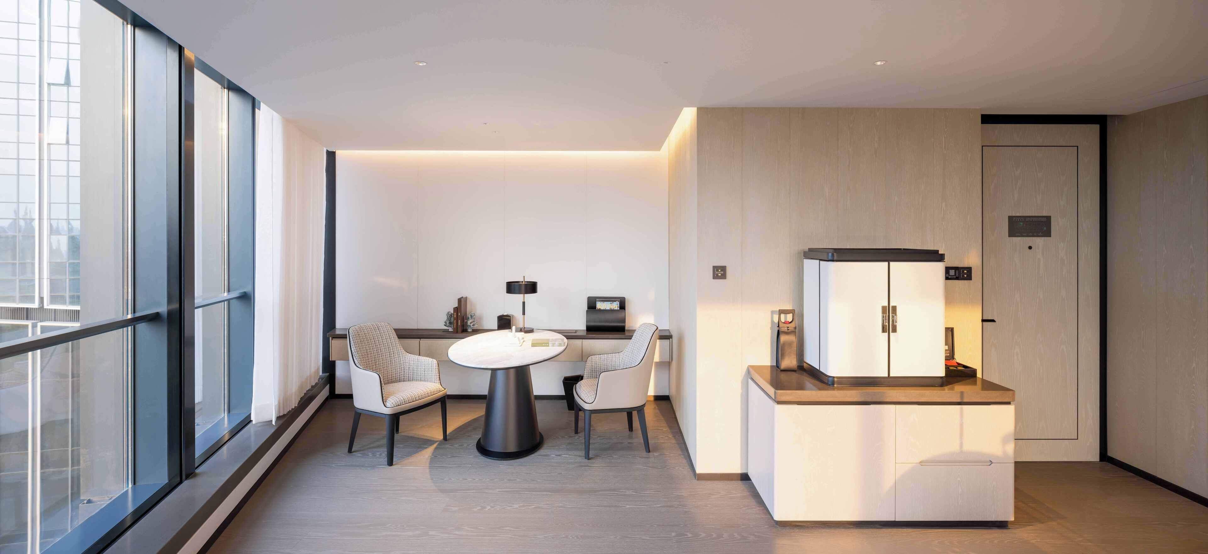 A modern hotel room with a large window, a round dining table with chairs, a minibar, and a wooden cabinet.