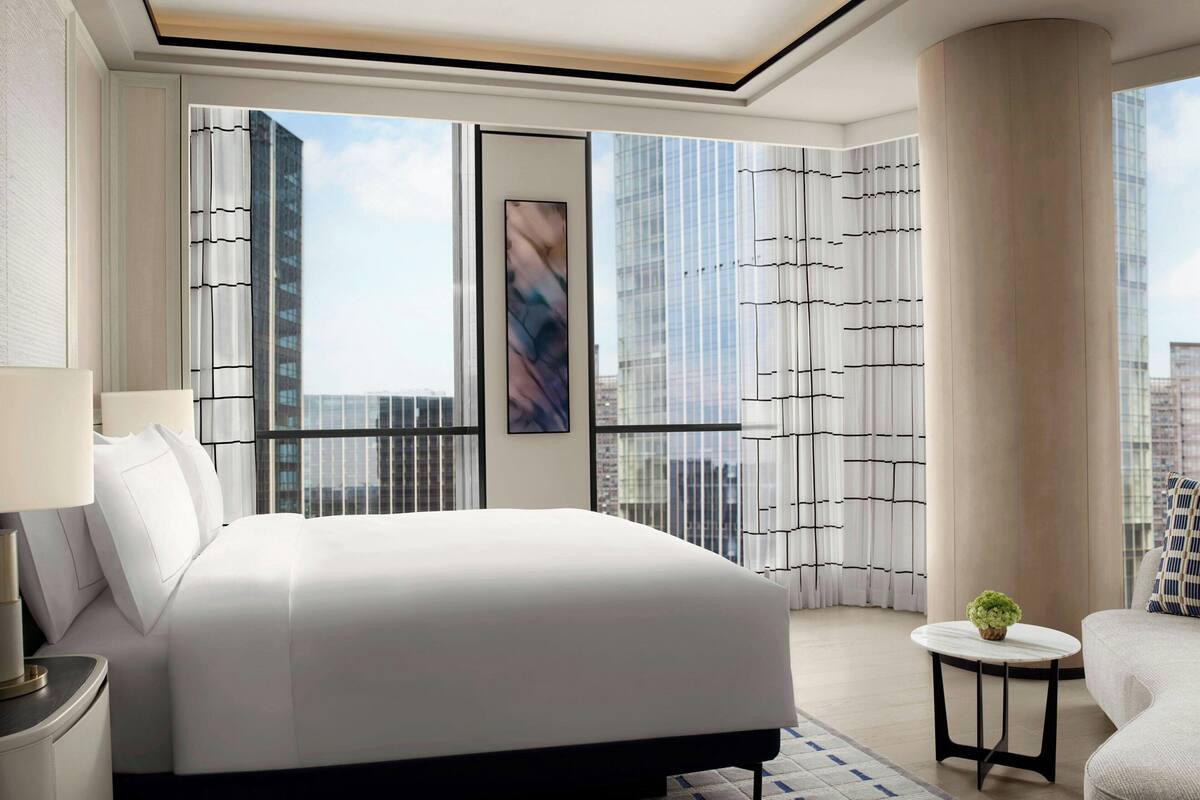 A modern hotel room with a large bed, a small round table with a plant, and a view of tall buildings through floor-to-ceiling windows.