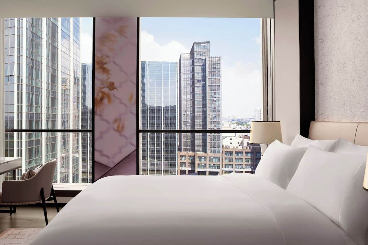 A modern hotel room with a large bed, a chair, and a view of city buildings through large windows.