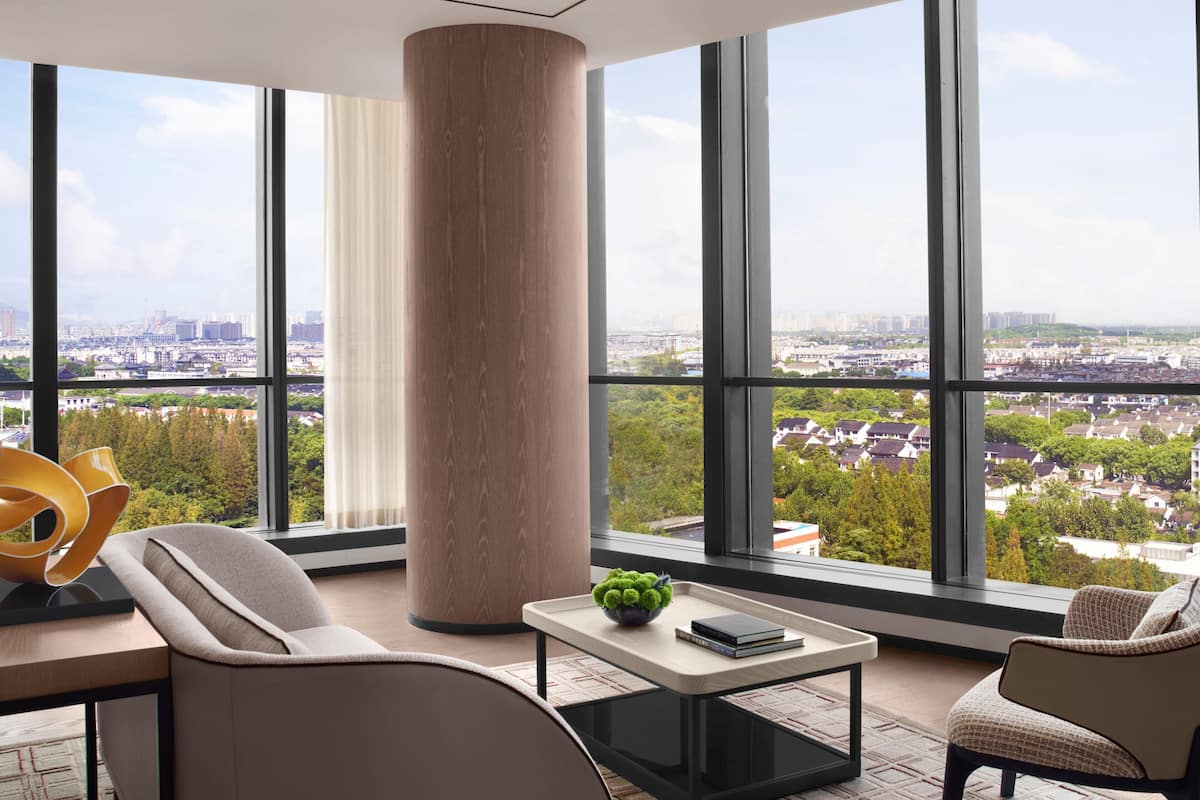 A modern hotel room with large windows offering a city view, a central column, and contemporary furniture.