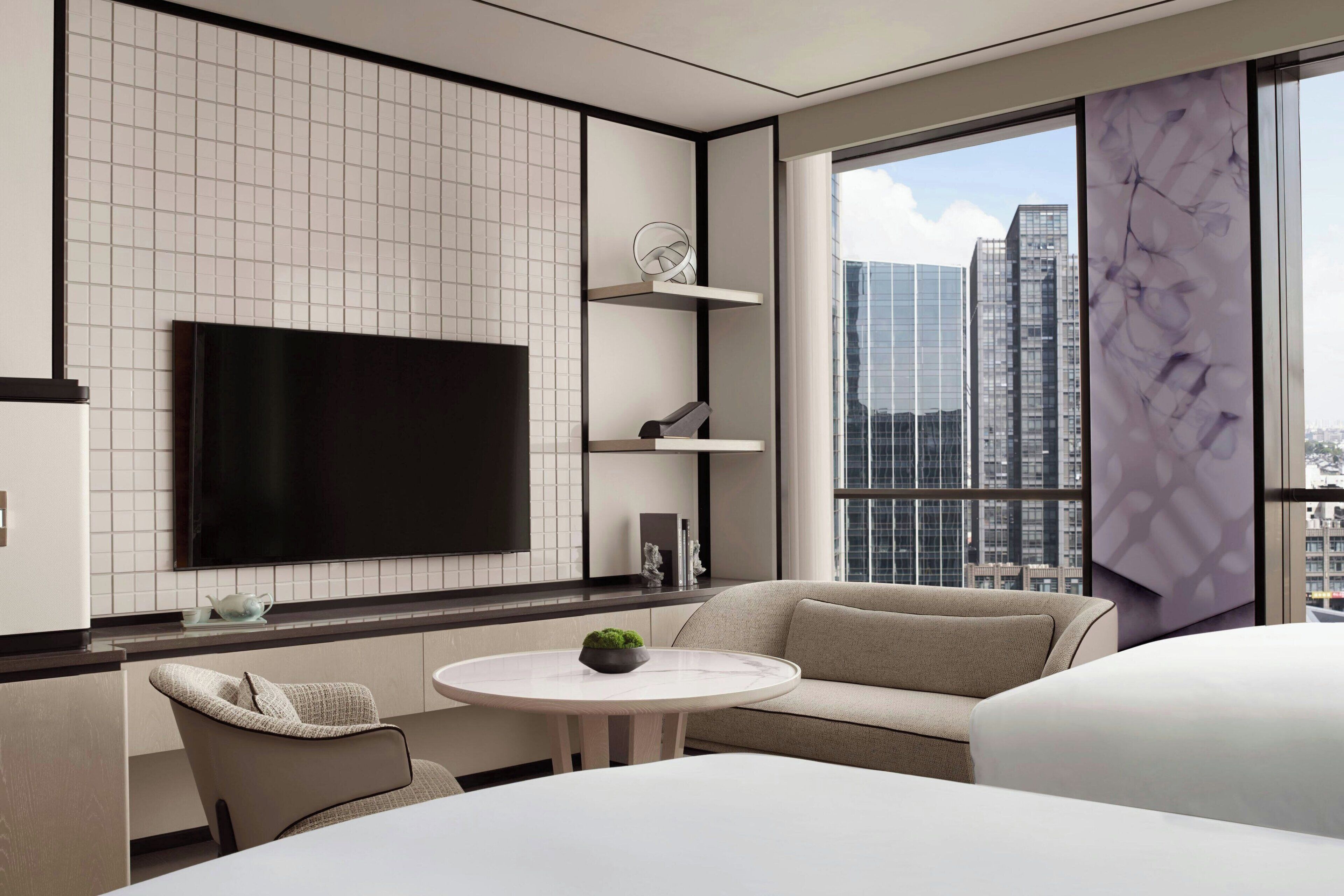 Quarto de hotel moderno com uma TV de tela plana grande fixada na parede, uma mesa de jantar redonda com uma pequena planta, um sofá e vista para a cidade através de grandes janelas.