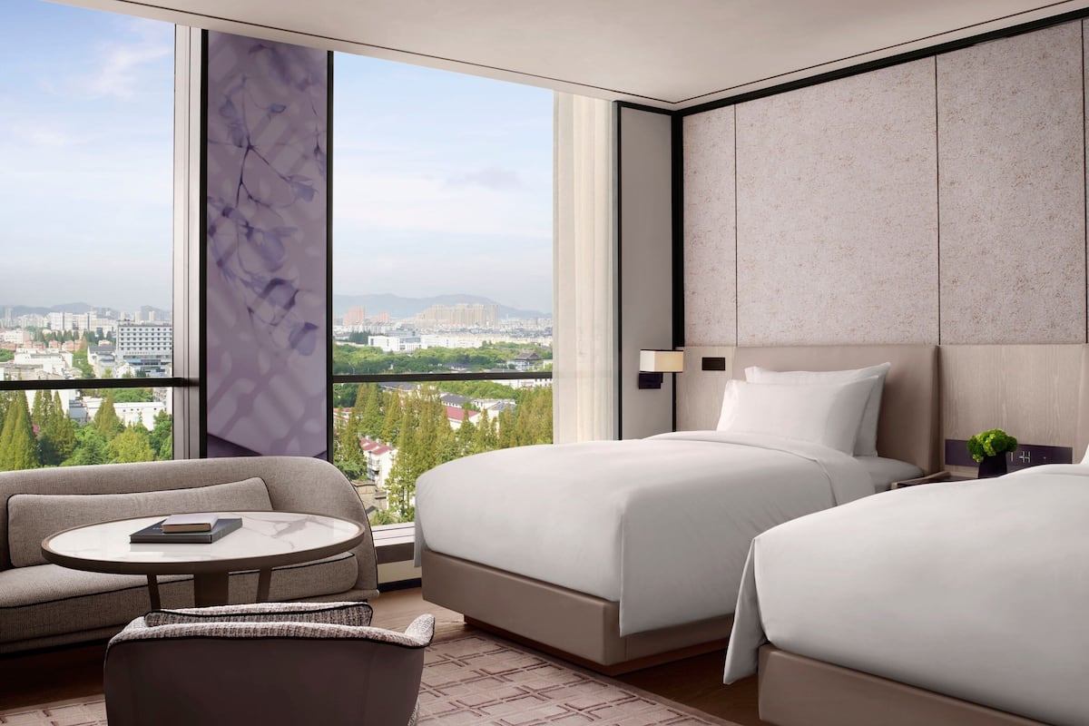 A hotel room with two beds, a sofa, a round table, and a large window offering a city view.