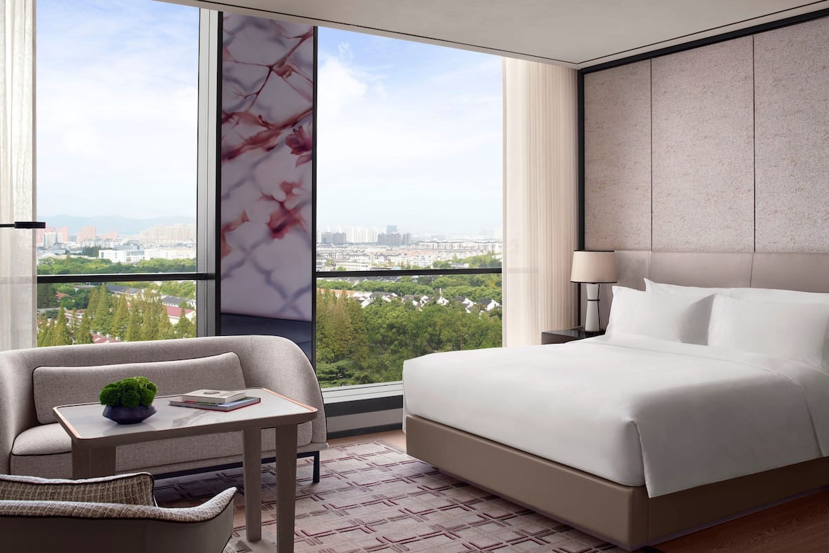 A modern hotel room with a large bed, a seating area with a table, and a view of a cityscape through large windows.