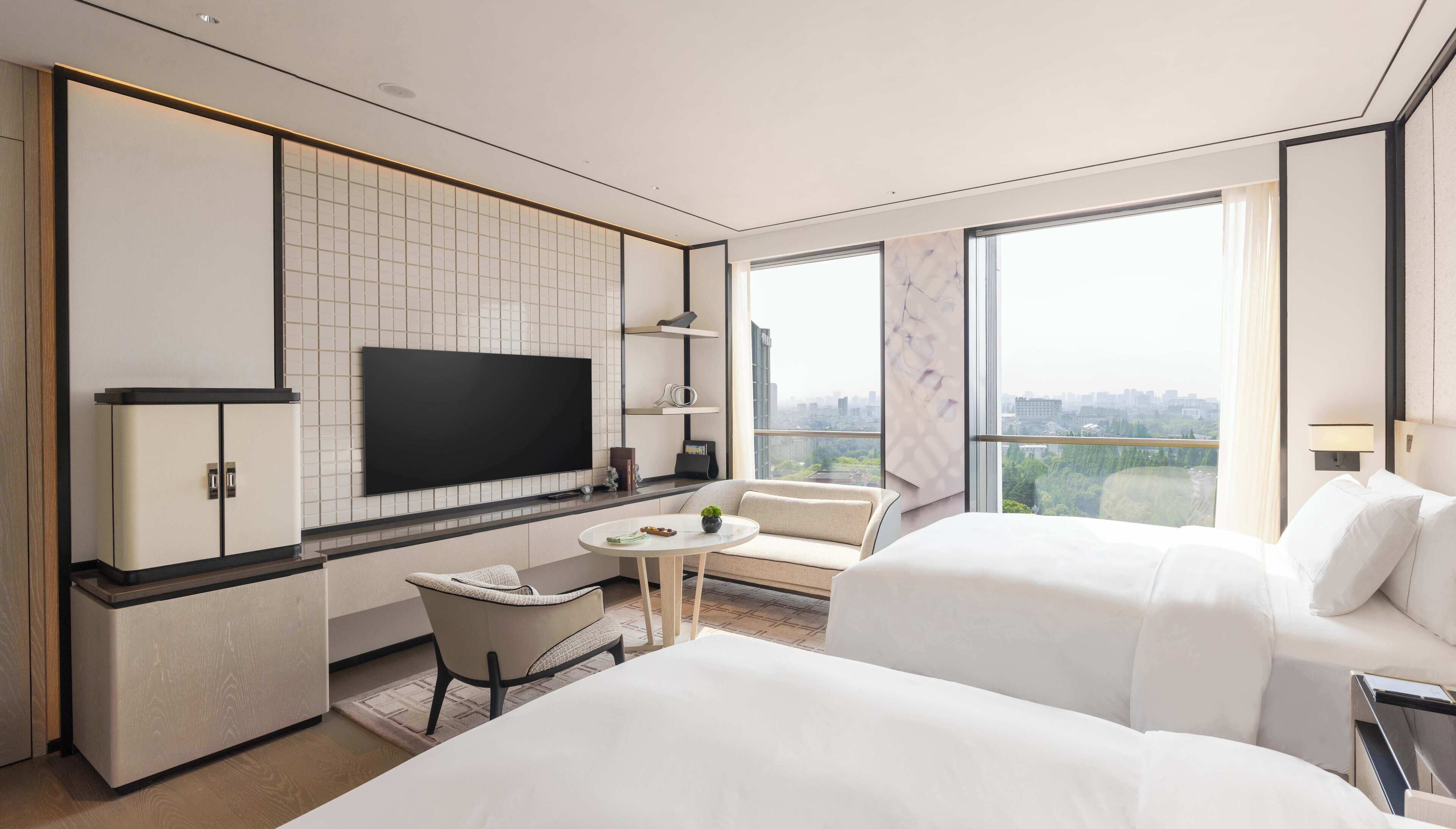 A modern hotel room with a large bed, a sofa, a small table, a TV, and a city view.