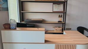 Un mostrador de recepción con acabados en madera clara, un panel blanco y un estante pequeño con un certificado enmarcado y figurillas.