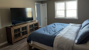Un dormitorio con una cama, un televisor sobre un mueble de madera, una ventana con persianas y una puerta.