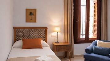 A hotel room with a bed, a bedside table with a lamp, a blue armchair, and a window with curtains.