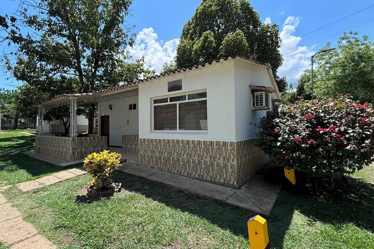 Un pequeño edificio de una sola planta con fachada de tejas, un porche cubierto y un jardín con un parterre amarillo y un arbusto de flores rosas.
