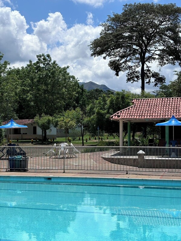 A swimming pool with a tiled deck, surrounded by a fence. There are two blue umbrellas providing shade.