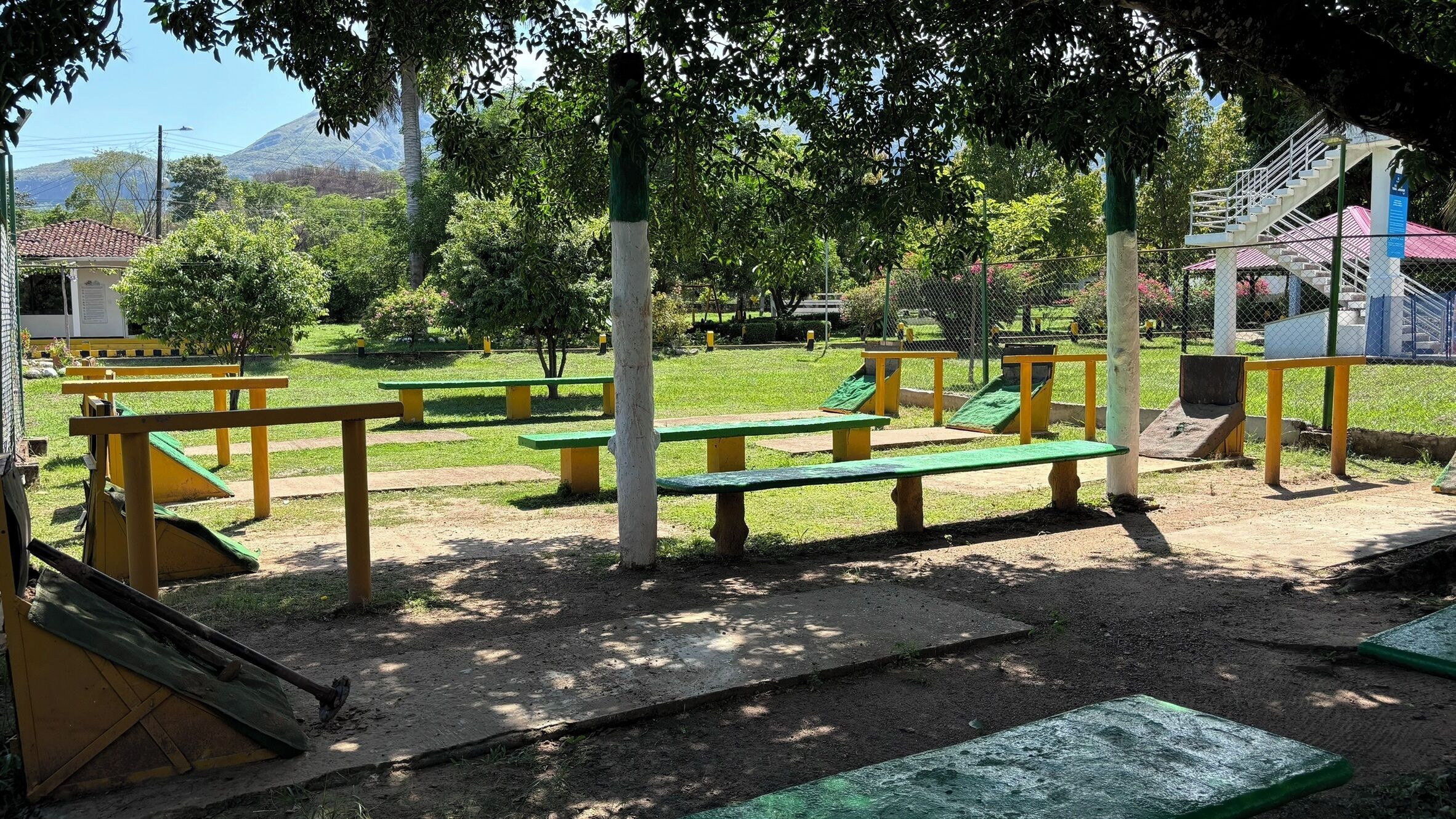 An outdoor playground with green and yellow equipment, including slides and swings, surrounded by trees and a view of a mountain in the background.