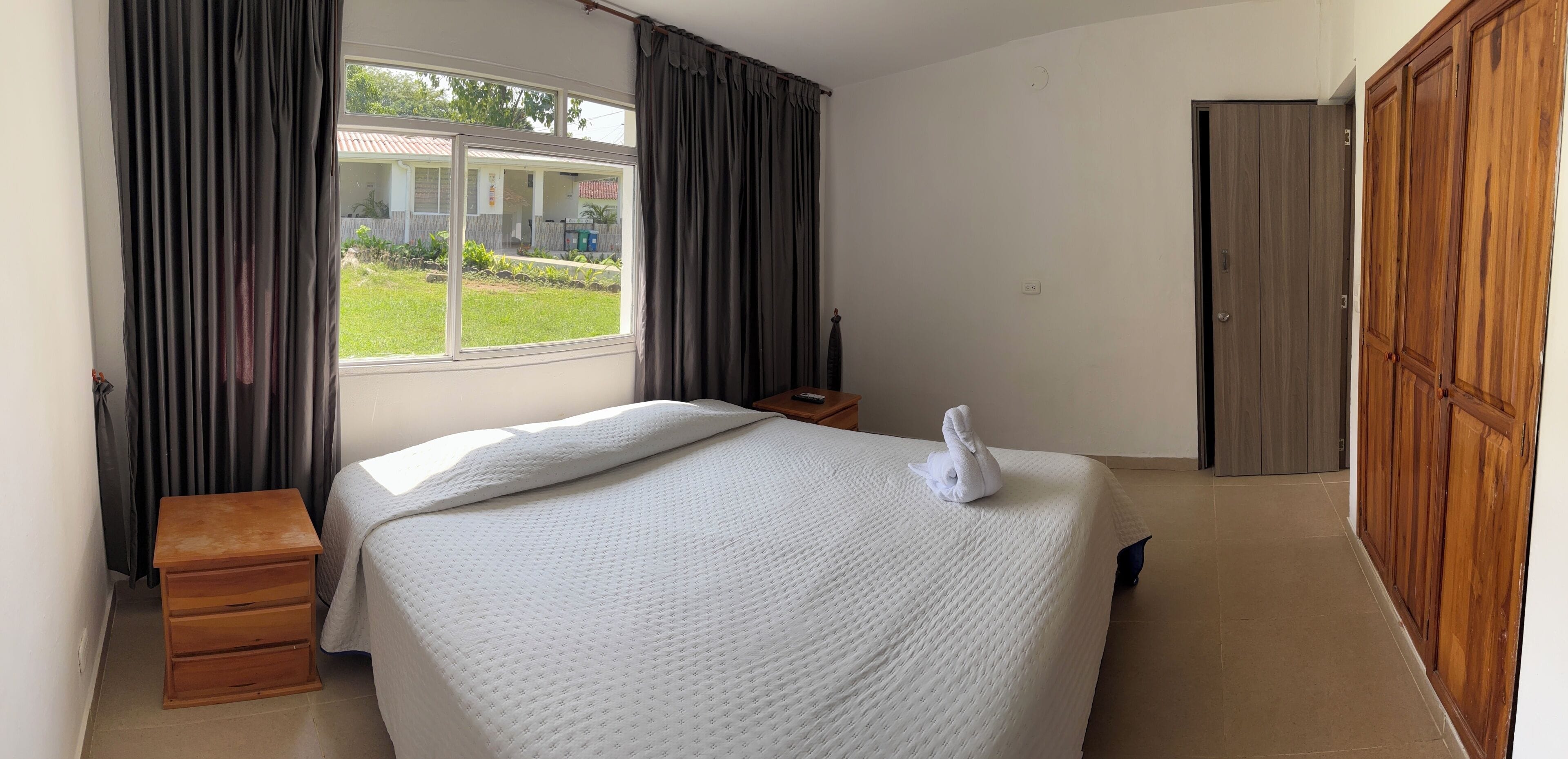 A hotel room with a bed, a wooden nightstand, a window with curtains, a wardrobe, and a view of the outside.