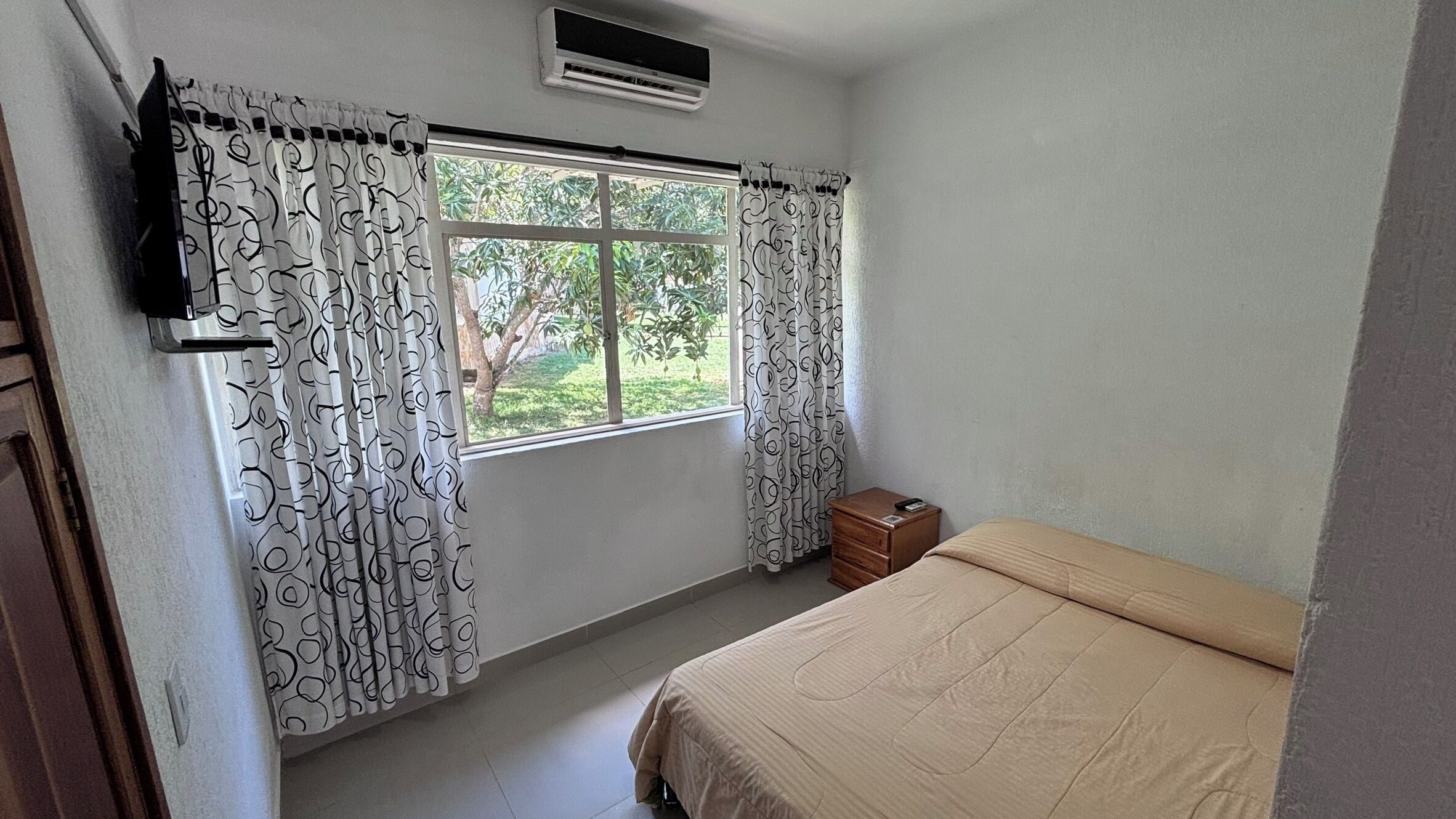 Un dormitorio con cama, ventana con cortinas, mesita de noche de madera y aire acondicionado.