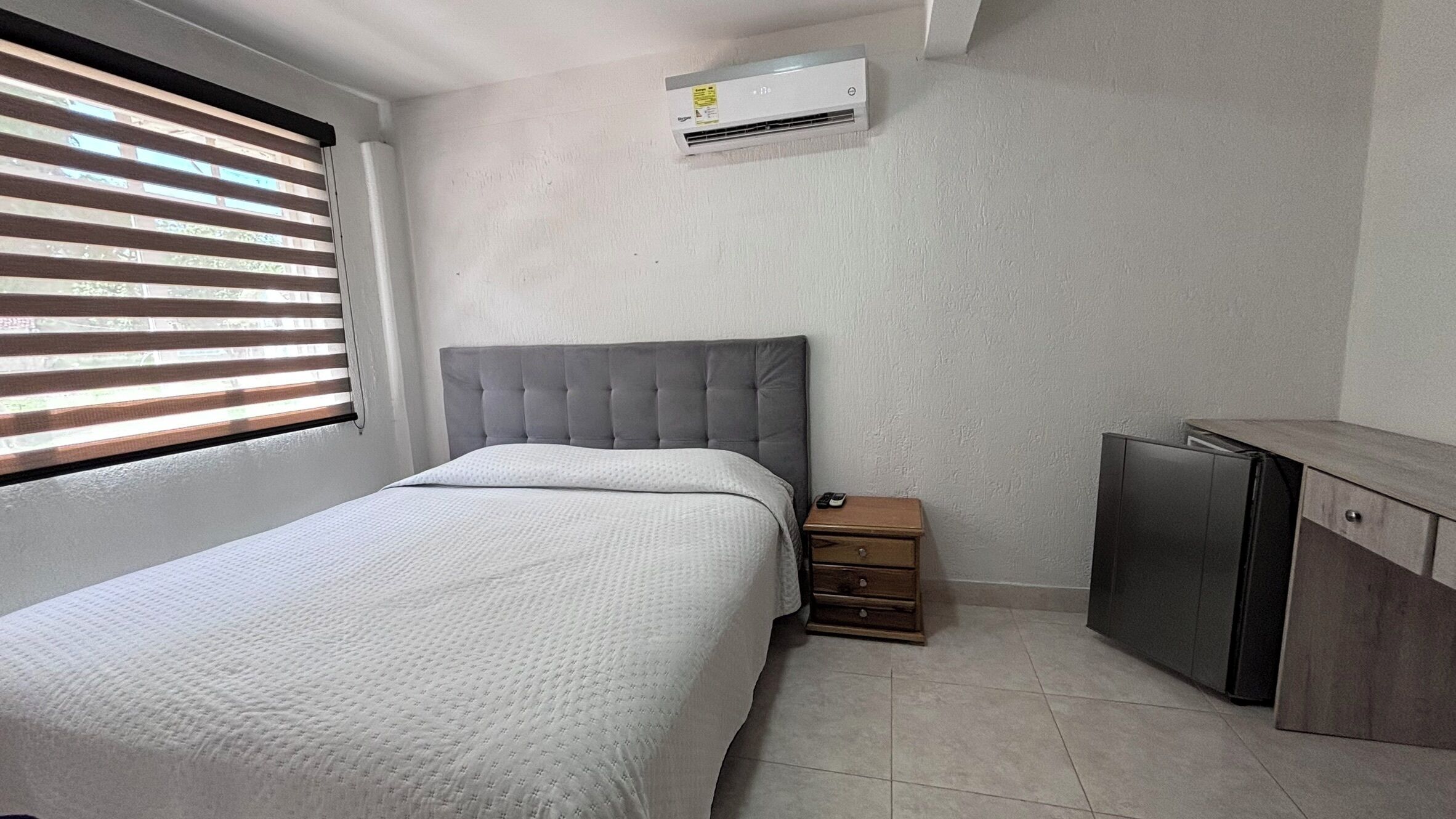 A bedroom with a bed, a nightstand, a small cabinet, and an air conditioning unit on the wall.