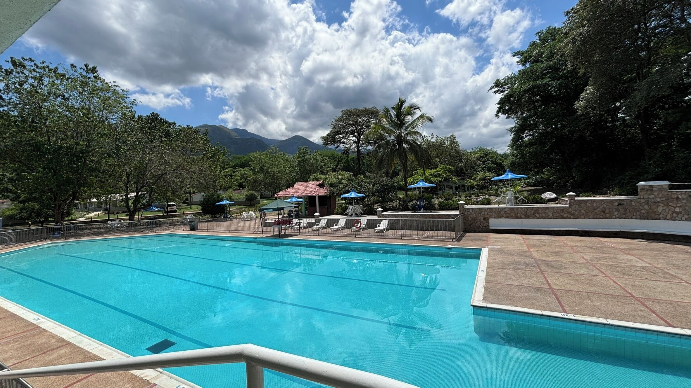 Una piscina con sillas de descanso y sombrillas, rodeada de árboles y una montaña al fondo.