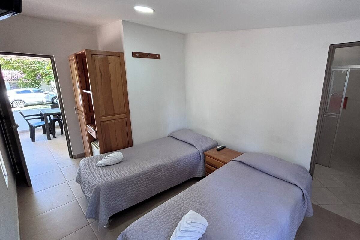 Una habitación pequeña y ordenada con dos camas individuales, un armario de madera, una mesita y una puerta.