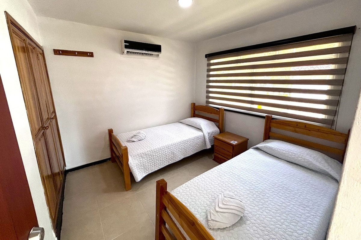 Una habitación pequeña y limpia con dos camas de madera, una ventana con persianas, un armario de madera y un equipo de aire acondicionado en la pared.