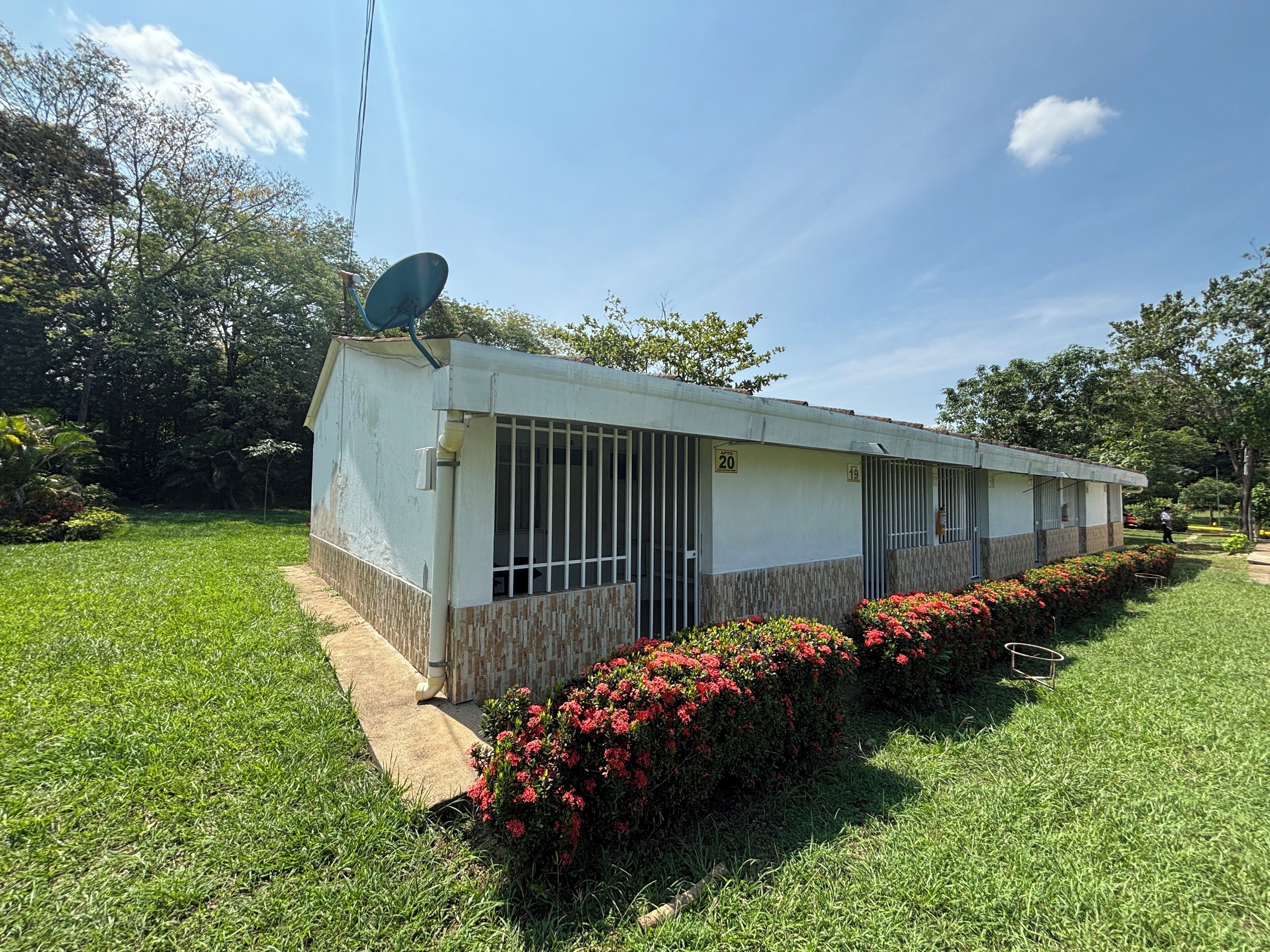 Edificio de una planta con antena parabólica, puertas numeradas y un jardín bien cuidado con flores rojas.