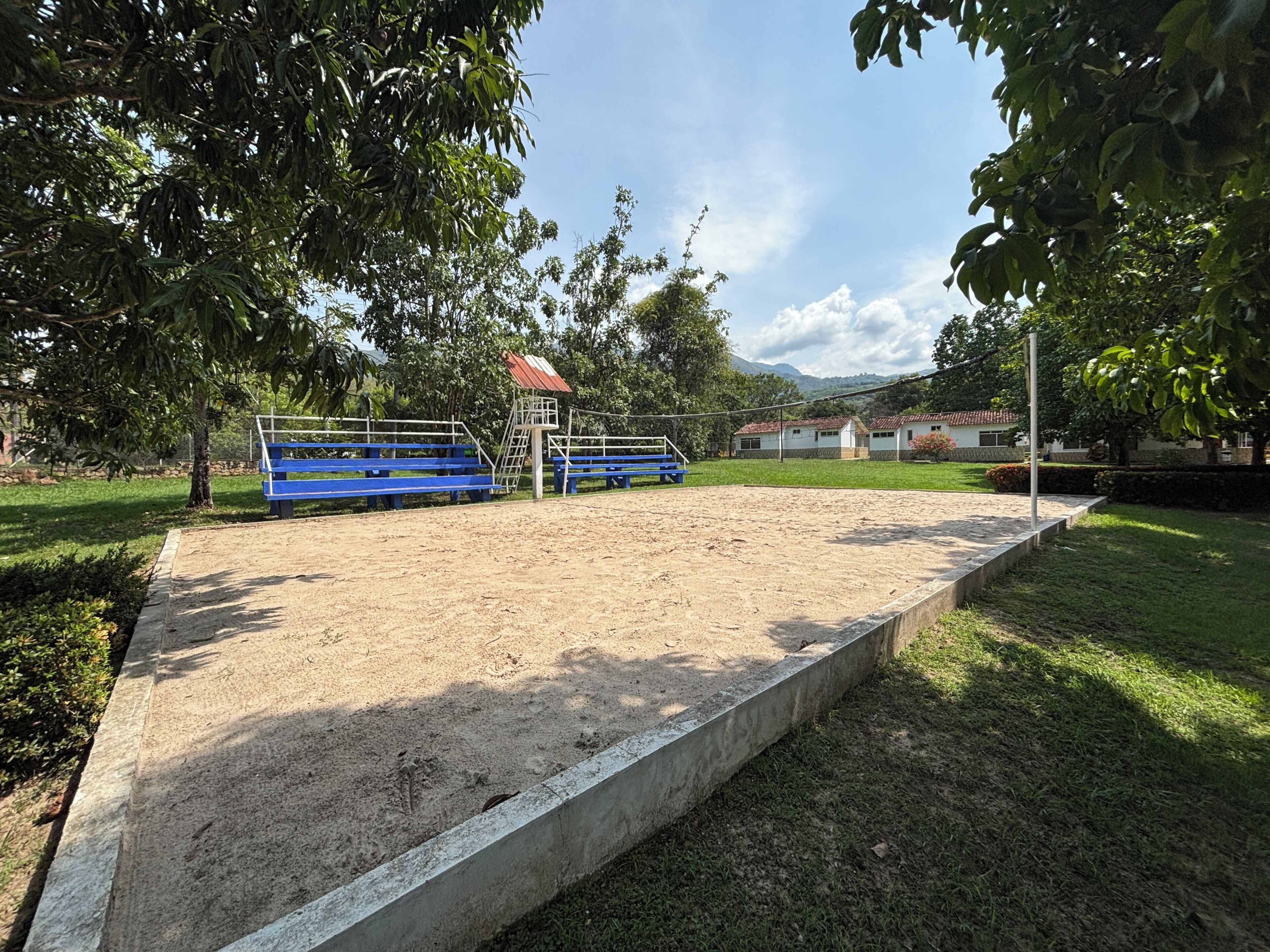 A sandy sports field with blue bleachers, a small shelter, and surrounding greenery.