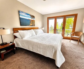 A bedroom with a large bed, a nightstand, a lamp, a chair, and a balcony with a view of trees.