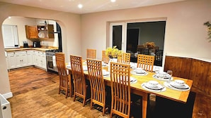 On aperçoit un coin repas avec une table en bois dressée pour quatre personnes, entourée de chaises en bois. Au fond, on distingue une cuisine avec des placards blancs et un fourneau.