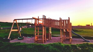 Une structure de jeux en bois, avec des balançoires et un toboggan, se dressant sur fond de ciel au coucher du soleil.
