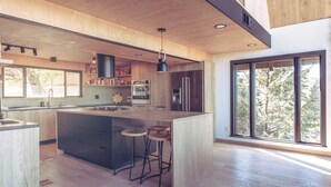 A modern kitchen with wooden cabinetry, a large island with stools, stainless steel appliances, and large windows offering a view of trees. - Breathtaking Designer A-Frame In The High Desert (Mountain Center)