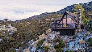 A modern house with a large glass facade, situated on a rocky hillside with a view of a winding road and mountains in the background. - Breathtaking Designer A-Frame In The High Desert (Mountain Center)