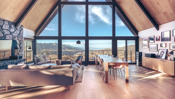 A spacious living area with a large window offering a mountain view, a cozy seating area, and a wooden dining table. - Breathtaking Designer A-Frame In The High Desert (Mountain Center)