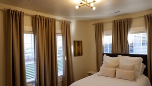 Een slaapkamer met een groot bed, beige gordijnen, een plafondlamp en een raam met zonwering.