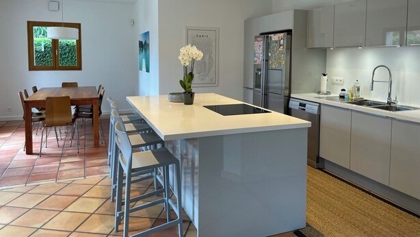 A modern kitchen with a central island, a dining area with a table and chairs, and a view of greenery through a window.