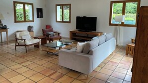 A living room with a sofa, armchair, and TV. There are windows and a door.