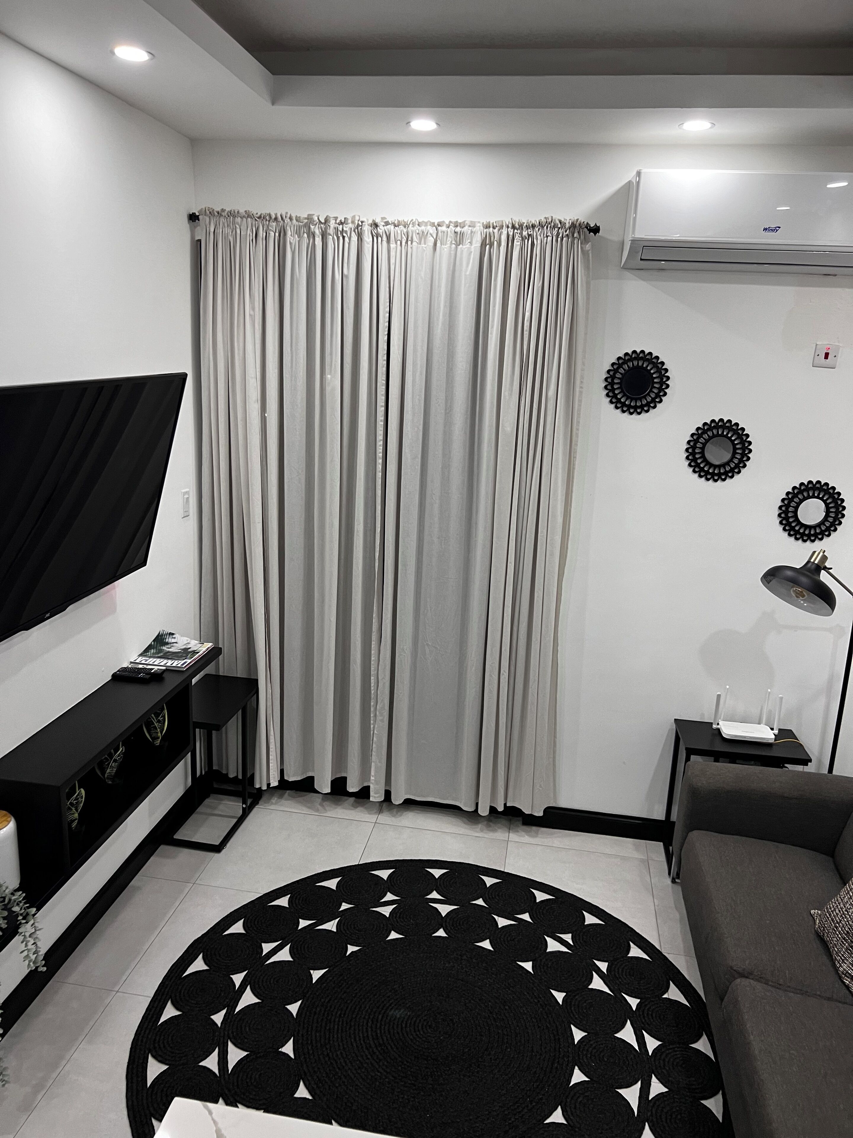 A modern living room with a flat-screen TV, a black console table, a grey sofa, a black and white patterned rug, and a grey curtain.