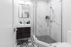 A modern bathroom with a glass-enclosed shower, a white sink with dark drawers, and a marble backsplash.