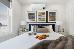 A hotel room with a large bed, framed pictures on the wall, a nightstand with a lamp, and a window with a view. - Parking 3BR2Bth, Cambridge/Boston, W/D, Subway,A/C (Somerville)
