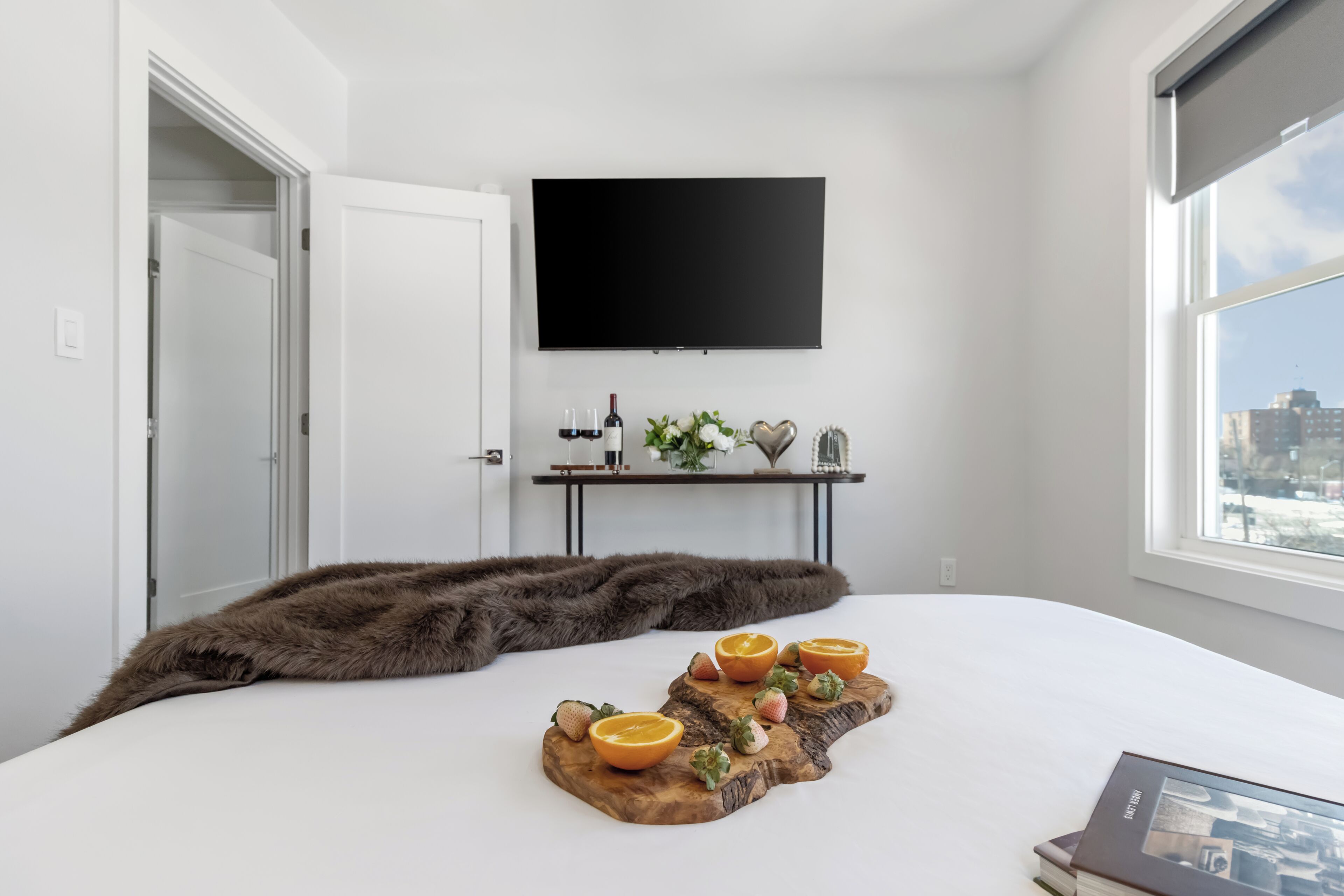 Un dormitorio con cama, televisor montado en la pared, ventana con vista a la ciudad y mesita de noche con planta y decoración en forma de corazón.