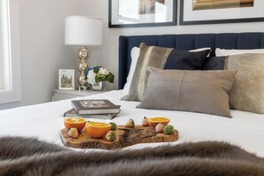 A neatly made bed with a wooden tray holding fruit, a lamp, and a framed picture on the nightstand.