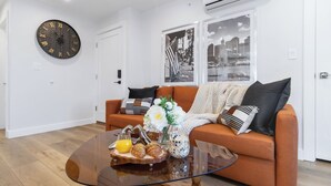 Un salon moderne avec une table basse ronde, un canapé orange, des coussins décoratifs, une horloge murale et des tableaux encadrés représentant des paysages urbains.