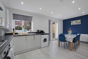 A modern kitchen with a built-in washing machine, a dining area with blue chairs, and a large window.
