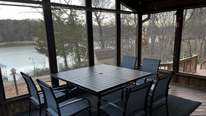 Eine überdachte Terrasse im Freien mit Tisch und Stühlen, mit Blick auf einen See und Bäume.