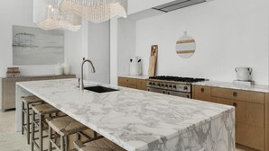 A modern kitchen with a large marble island, wooden stools, a gas stove, and a hanging light fixture.