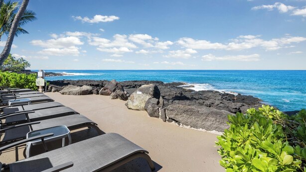 A beachside view with lounge chairs, a rocky shoreline, and a clear blue sky. - Kona Makai #5203 Ocean View , 
A Hawaiian Paradise (Kailua-Kona)