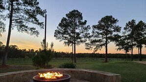 A stone fire pit with a fire in the center, surrounded by a stone wall, set in a grassy outdoor area with trees in the background. - The Vine - Elegant "Wild Rose" Cottage on Vineyard with Private Lake, Sleeps 4 (New Ulm)