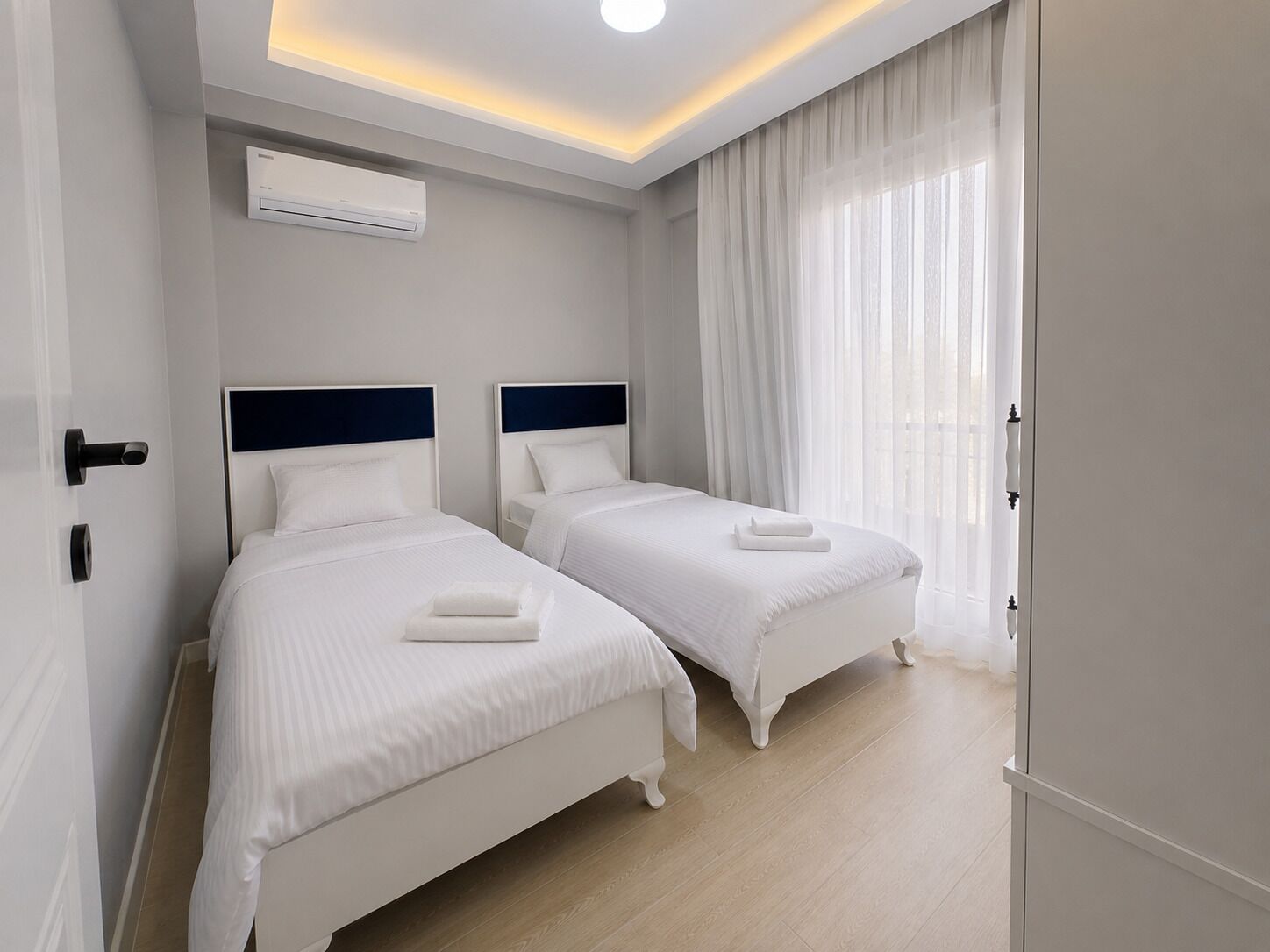 A hotel room with two beds, a ceiling light, an air conditioning unit, and a large window with sheer curtains.